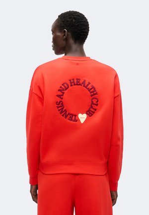 Personne aux cheveux courts portant un sweat-shirt rouge avec le texte "Tennis and Health Club" en cercle et un petit cœur dans le dos.