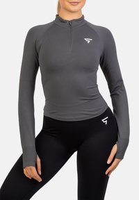 Szary, długi rękaw, sportowy top z zamkiem na ćwierć, z wysokim kołnierzem i subtelnym logo; połączony z czarnymi legginsami. Gładka tekstura.