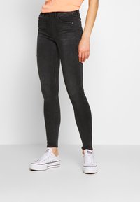 Pantalones vaqueros negros ajustados de denim, con una cintura alta, diseño de cinco bolsillos y un aspecto desgastado. Combinados con zapatillas blancas.