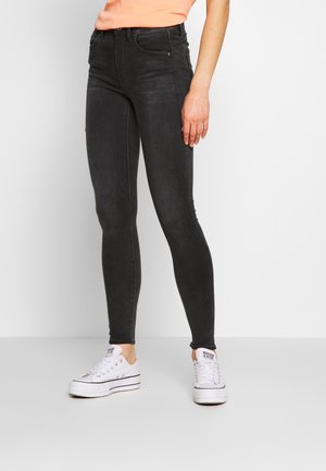 Pantalones vaqueros negros ajustados de denim, con una cintura alta, diseño de cinco bolsillos y un aspecto desgastado. Combinados con zapatillas blancas.