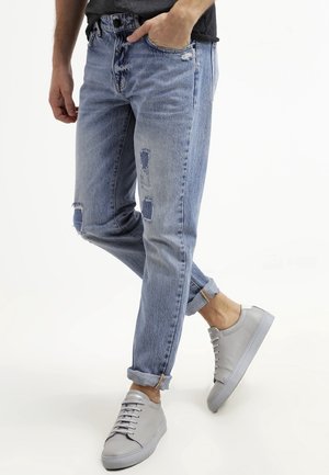 Mężczyzna stojący z rękami w kieszeniach, ubrany w jasnoniebieskie postrzępione dżinsy podwinięte przy nogawkach oraz jasnoszare trampki na białym tle.