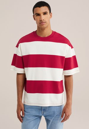 Jeune homme portant un t-shirt oversize à rayures horizontales rouges et blanches et un jean bleu clair, debout devant un fond uni.