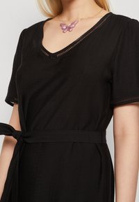 Musta mekko lyhyillä hihoilla, v-pääntiellä ja sidottavalla vyötäröllä. Sisältää teksturoitua kangasta ja ohut aukko-detaili pääntien ympärillä.