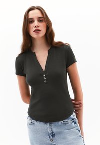 Donkergroene short-sleeve top met een V-hals, voorzien van vier zilveren knoopaccenten en een geribbelde textuur, gecombineerd met lichtblauwe jeans.