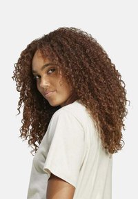 Cabello rizado hasta los hombros en un tono marrón cálido. Lleva una camisa de color claro y de manga corta. Fondo neutral.