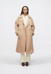 Μπεζ καμπαρντίνα με φαρδύ γιακά, διπλή σειρά κουμπιών και πλαϊνές τσέπες. Φορεμένη πάνω από ανοιχτόχρωμο τοπ και παντελόνια με φαρδιά μπατζάκια.