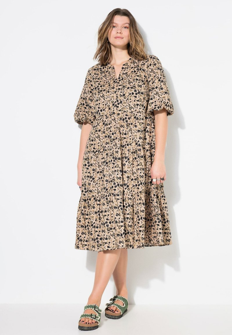 Korte mouwen, jurk tot de knie in tan met een zwart dierenprint. Bevat een knoopsluiting op de kraag en een geplooide rok. Gemaakt in combinatie met groene platform sandalen.