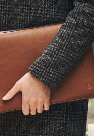 Brun läder laptopfodral som hålls i en hand, med en slät yta och en logodetalj. Bakgrunden är en texturerad mörk kappa.