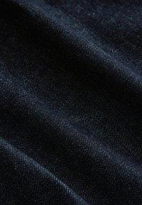 Tissu en denim indigo foncé avec texture visible de tissage diagonal et plis doux créant des variations de lumière et d'ombre.