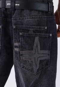 Los vaqueros de mezclilla gris oscuro presentan un diseño de bolsillo distintivo con un patrón de estrella en relieve y detalles sutiles de costura. Se puede ver un cinturón de cuero negro.