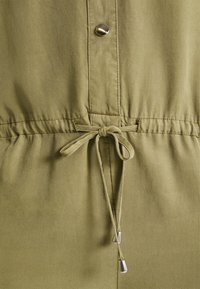 Tissu vert olive avec une taille à cordon de serrage noué et un bouton en argent au-dessus de la couture sur un vêtement.