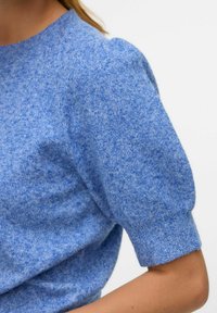 Pull en maille bleu avec manches bouffantes courtes, présentant un tissu texturé et un col ras du cou. La couleur a un subtil effet chiné.