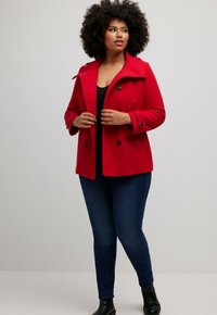 Cappotto rosso doppiopetto con colletto a rever, bottoni neri e texture morbida, abbinato a jeans blu e stivali neri alla caviglia.
