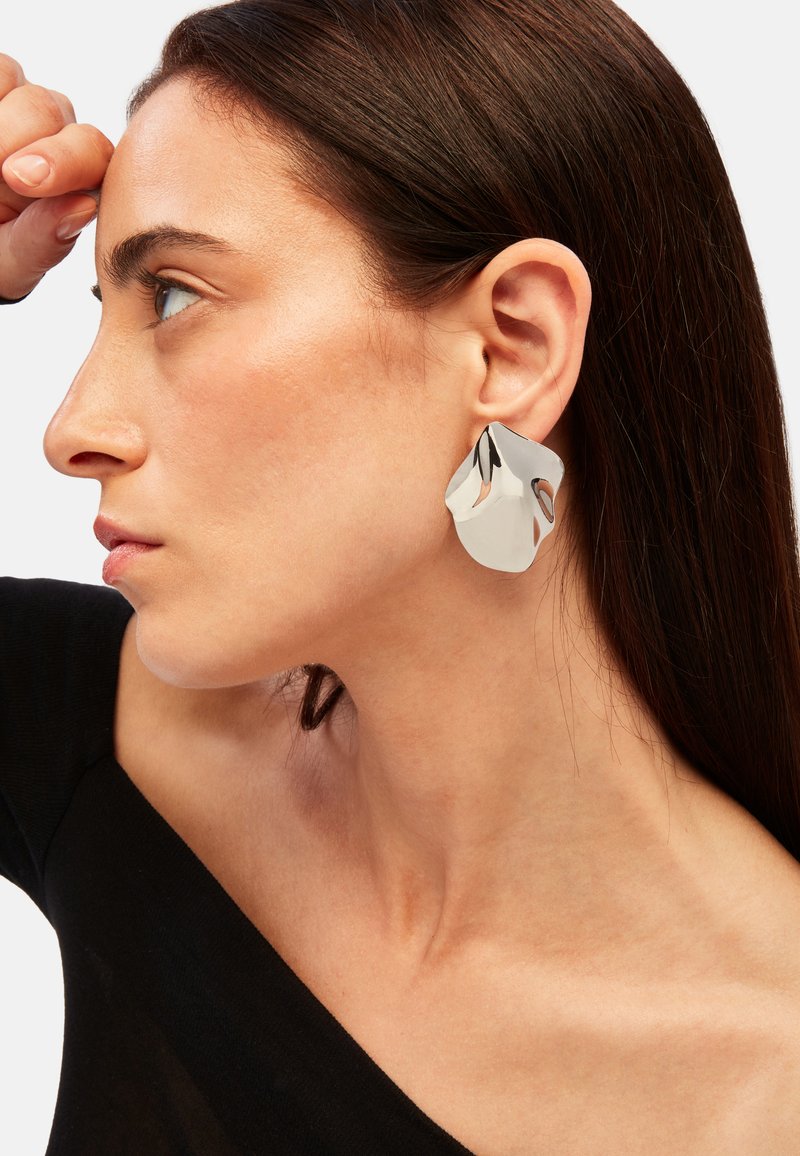 Stora, abstrakta örhängen i silver med en vikt design och utskärningsdetaljer. Bärs på vänster öra och visar en polerad, spegelblanka yta.