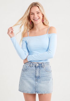 Smilende kvinde med langt blondt hår bærer lys blå off-shoulder top, denim nederdel og farverige kæde-øreringe, holder hår med den ene hånd.