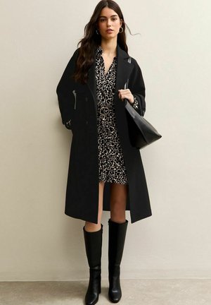 Vrouw draagt een lange zwarte jas over een jurk met luipaardprint, zwarte kniehoge laarzen en houdt een zwarte schoudertas vast tegen een effen achtergrond.