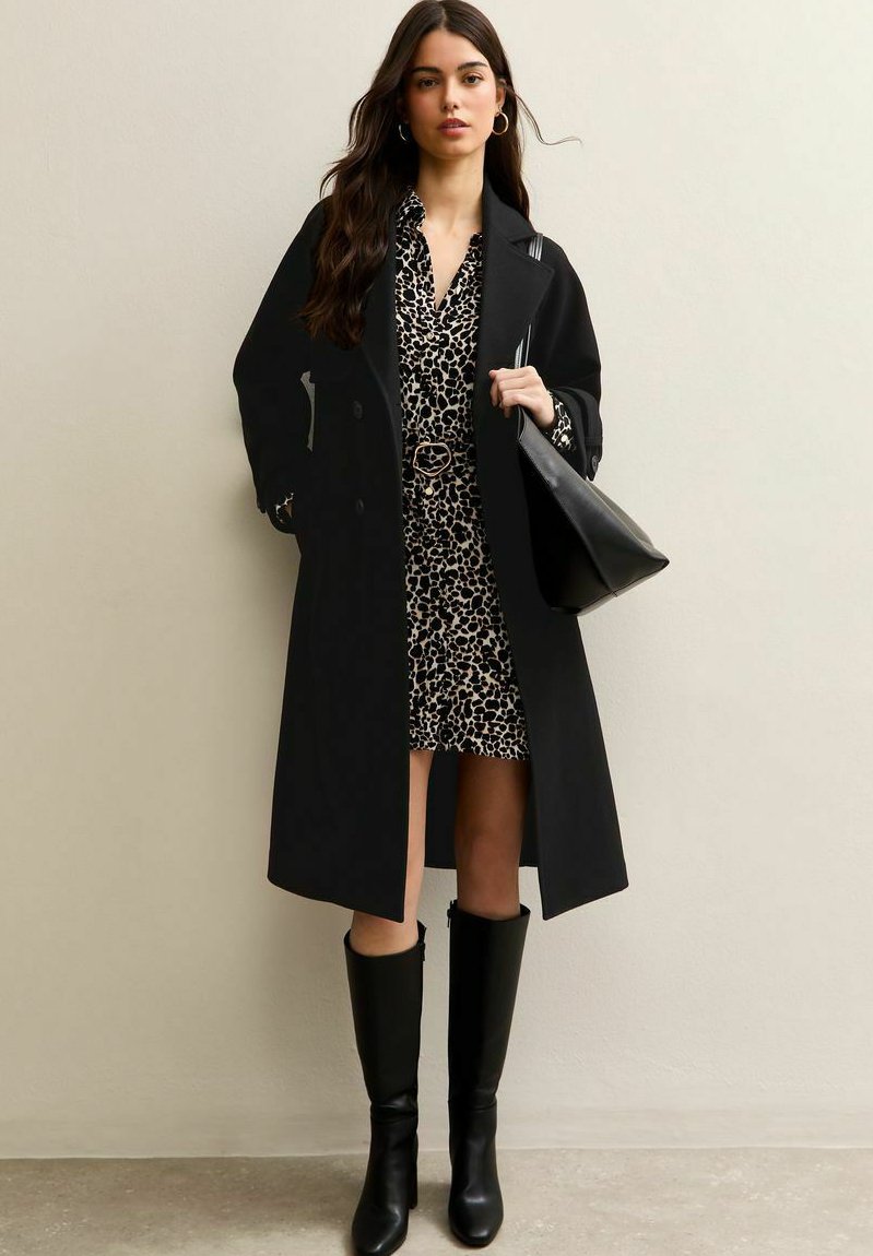 Vrouw draagt een lange zwarte jas over een jurk met luipaardprint, zwarte kniehoge laarzen en houdt een zwarte schoudertas vast tegen een effen achtergrond.