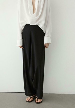 Persona che indossa pantaloni neri larghi e ampi, camicetta bianca a maniche lunghe con scollo drappeggiato e sandali neri open-toe su un pavimento grigio.