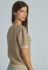 Maglione a maniche corte in beige con un motivo a trama. Presenta un colletto ondulato e polsini a coste. Tessuto morbido e leggero.