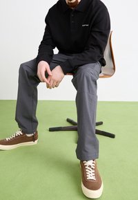 Man zit op houten stoel met zwart langemouwen shirt, grijze broek en bruine sneakers op groene vloerbedekking.
