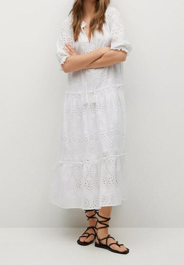 Femme portant une robe midi blanche à œillets avec des manches trois-quarts et des sandales noires lacées, debout les bras croisés.