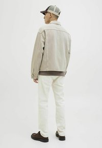 Giacca beige chiaro con una vestibilità rilassata, maglietta marrone, jeans bianchi e scarpe marroni scure. Il cappellino ha un retro in rete e un visiera scura.