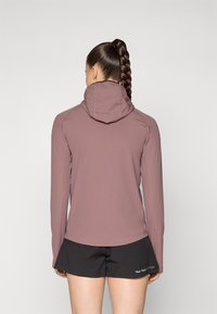 Hoodie ajusté à manches longues en mauve avec capuche, comportant des coutures contrastées. Associé à un short noir, montrant une texture lisse et extensible.
