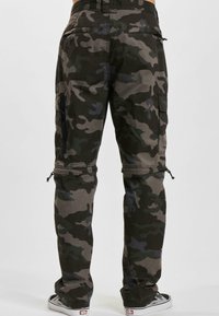 Man som bär mörkgrön och grå kamouflagebyxor och svartvita sneakers, står med raka ben mot en enfärgad bakgrund.