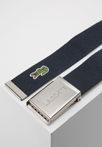 Navy woven belt with a metal silver buckle, featuring an embossed brand name and a green crocodile logo embroidered on the strap.