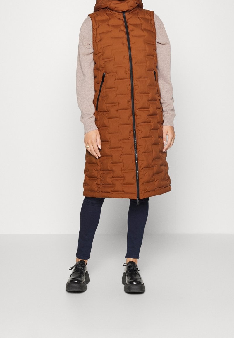 Personne portant un long gilet matelassé rouille-orange avec fermeture éclair à l'avant, un pull beige, un pantalon skinny foncé et des chaussures à plateforme noires à lacets.