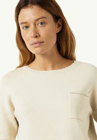 Sweter w kolorze kremowym, wykonany z miękkiej tkaniny, z okrągłym dekoltem, prążkowanym wykończeniem i małą kieszonką na piersi z kontrastowym szwem.