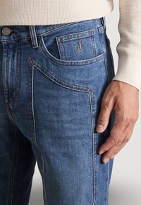 Los jeans de mezclilla azul presentan un diseño ajustado con estilo de cinco bolsillos. La tela tiene una ligera textura y hay un sutil logo de la marca en el bolsillo delantero.