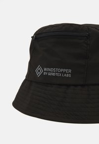 Svart bøttehatt laget av Windstopper-stoff, med en glidelås-lomme på siden og merket logo i grått. Teksturet og vannavvisende.