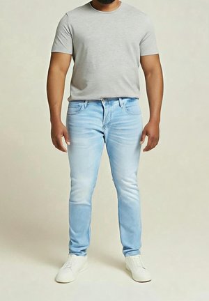 Hombre con camiseta de manga corta gris claro, vaqueros azul claro y zapatillas blancas, de pie frente a un fondo beige liso.