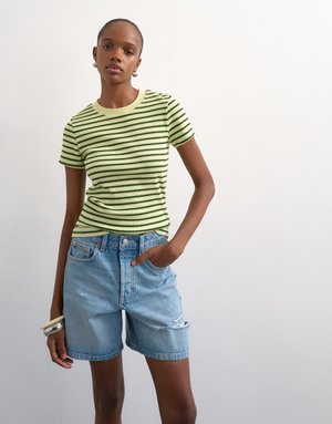 Jeune femme portant un t-shirt moulant à rayures vert clair et noir avec un short en denim bleu clair déchiré, debout devant un fond blanc uni.