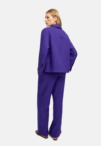 Fioletowy, teksturowany dwuczęściowy strój składający się z kurtki z kołnierzykiem i spodni o szerokich nogawkach, z guzikami na rękawach kurtki.
