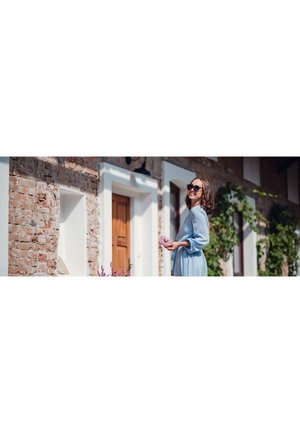 Light blue, flowy dress with ruffled sleeves; person holding a pink wallet; brick wall and wooden door in background; sunny day.