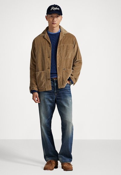 Mann iført en marineblå Ralph-caps, lys cordfløyelsjakke, blå kabelstrikket genser, falmede jeans og brune semsko som står mot en enkel bakgrunn.