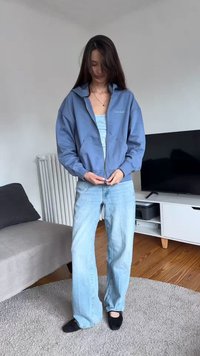 Ung kvinde med langt mørkt hår, der lynlåser en blå Calvin Klein hættetrøje, iført lyseblå jeans og sorte hjemmesko i en stue.