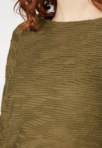 Top verde oliva realizzato in tessuto testurizzato con modelli superficiali ondulati. Collo tondo, caratterizzato da sottili variazioni nella texture e nella profondità.
