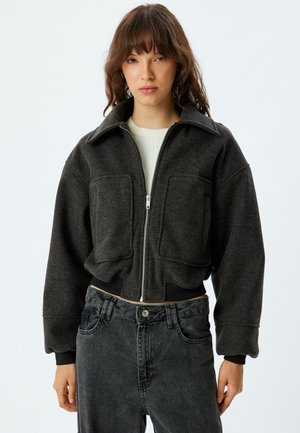 Gekürzte dunkelgraue Jacke mit großen Vorderaschen, einem Reißverschluss und einem spitzen Kragen. Getragen über einem hellen Oberteil und schwarzen Jeans.