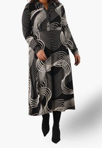 Lungo vestito nero con linee bianche ondulate, design con colletto, maniche lunghe e silhouette fluente. Indossato con collant neri aderenti e scarpe a punta.