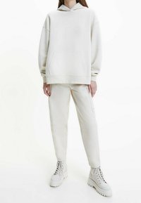 Sudadera beige clara con bolsillo canguro y capucha con cordones, combinada con pantalones ajustados a juego que cuentan con un bolsillo lateral y botas gruesas blancas.