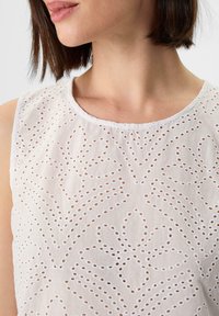 Biała bezrękawnikowa bluzka z misternym haftem w technice eyelet, z kwiatowymi i geometrycznymi wzorami, okrągłym dekoltem i lekkim materiałem.