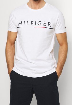 Mann trägt weißes T-Shirt mit kurzen Ärmeln und Schriftzug "HILFIGER" sowie farbige Akzente, kombiniert mit schwarzen Hosen, Hände in den Taschen.