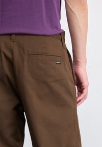 Bruine katoenen broek met een rechte snit, voorzien van een achterzak en een klein zwart label. De tailleband is zichtbaar boven de zak.