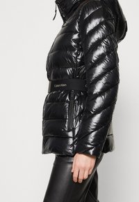Persona vistiendo una chaqueta acolchada negra brillante con un cinturón de marca Calvin Klein y pantalones de cuero negros, de pie contra un fondo claro y plano.