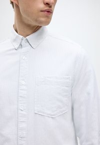 Λευκό πουκάμισο με λεπτές κάθετες ρίγες, γιακά με κουμπιά, τσέπη μπροστά και κανονικά κουμπιά. Το υλικό φαίνεται μαλακό με διακριτή υφή.