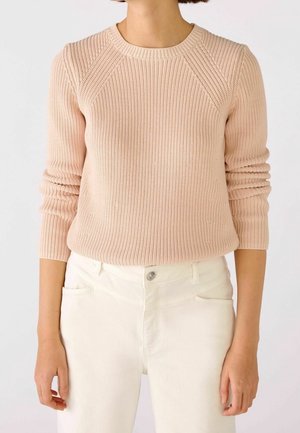 Femme portant un pull en maille côtelée beige clair à manches longues, associé à un pantalon taille haute blanc cassé.