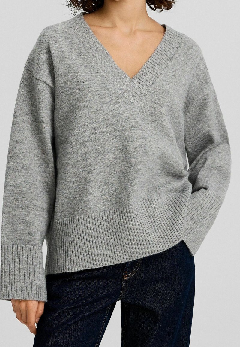 Personne portant un pull gris à col en V ample avec des poignets et un bas côtelés, associé à un jean bleu foncé, main dans la poche.